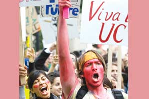 Students with face covered in red dye carrying sign protesting against funding cut at University California system.