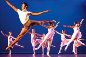 Lead male ballet dancer Gabriel with white T-shirt and short tight in efface derriere position in the air with seven female dancers dress in light pink ensembles dancing behind him. The stage is lid with deep sea blue color background.