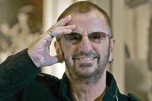 A portrait of Ringo Starr smiling with his right hand touching his face.