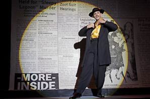 The character El Pachuco (played by Joel Mijares) stand in bright circular spot light on the stage with Zoot-Suit style black suit and hat with bright busy yellow-and-white patterned inner shirt. The background of the stage is a giant print of newspaper with story about the Zoot-Suit court hearing.