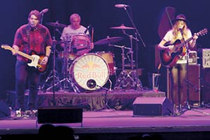 Nicole Vaughn singing while playing her guitar on the stage with her band base player and drummer under purple light.