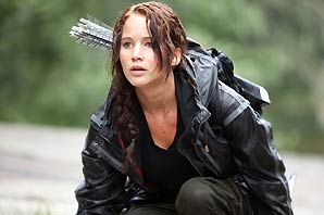 Actress Jennifer Lawrence in a scene of her portrayal of Katniss Everdeen where her messed up hair is tied up at the back of her head, she has bloody scratch wounds on her neck and chest, she wore a black vinyl jacket, black shirt, grey pants, with arrows in arrow holder on her back while she is in semi-squatting position in the woods.