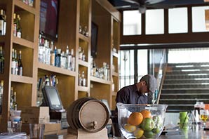 A mixologist working behind the counter of a restaurant. The counter is displayed with a table top size beer barrel, some fresh fruits in a big glass bowl, condiments, and glasses. On the back of the counter are shelves filled with hundred of bottles of alcoholic beverages, and three television sets.