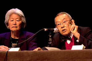 Dr. Donal Teruo Hata sharing testimonies in front of a microphone while Hatsuko Mary Higuchi watches on next to him.