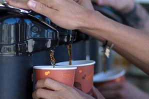 Three customers pressing on the handle of spouts of brewer containers to fill their cups with hot beverages.