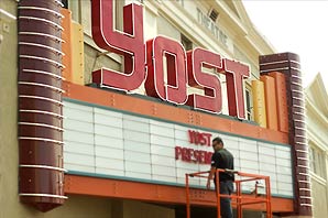 A worker on top of construction stand in front of big and colorful YOST sign outside Yost Theater placing sign which reads "YOST PRESENT…"