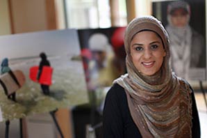 Photojournalist and author Sadaf Syed wearing a headscarf, in a room surrounded by photos of women in different settings, including surfing.