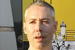 Closeup photo of Director Adam Yauch standing in front of the "'Gunning' For That #1 Spot" poster. The poster with yellow background and black text has black-and-white images of basketball players all holding basketball in their hands.