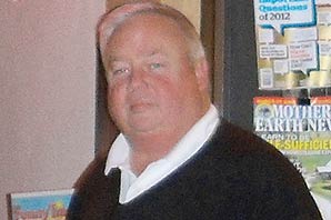 Owner of RAG-International-News-Stand, wearing a white shirt with dark color sweater, stand in front of his store with rags of magazines and newspapers by the side of the store.