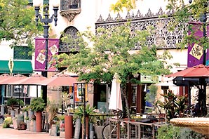 View outside of the Santora Building and stores along the same street; there are many sitting areas under red-peachy color big umbrellas and there are several trees and smaller plants of varying sizes beside the sitting areas as well as a ceramic water fountain on the sidewalk.