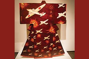 An art piece completely made of wood is a Japanese kimono with reddish brown background, a Japanese symbol, two Chinese/Japanese written characters and repetitions of WWII Japanese fighting jets and bomb blasts.