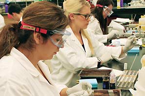 Several women in white lab coat and protective googles, wearing plastic gloves handing specimens.