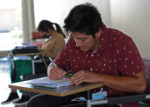 Primary subject is a maskless student seated, with pencil in hand and his mask on the chair arm. Student in foreground (masked) while seated with a laptop.