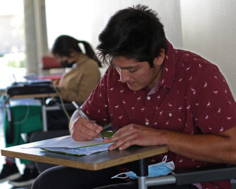 Primary subject is a maskless student seated, with pencil in hand and his mask on the chair arm. Student in foreground (masked) while seated with a laptop.