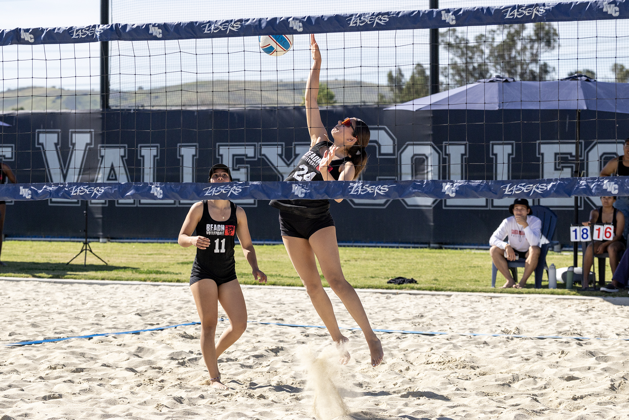 Female volleyball player jumps on sand court to block shot.