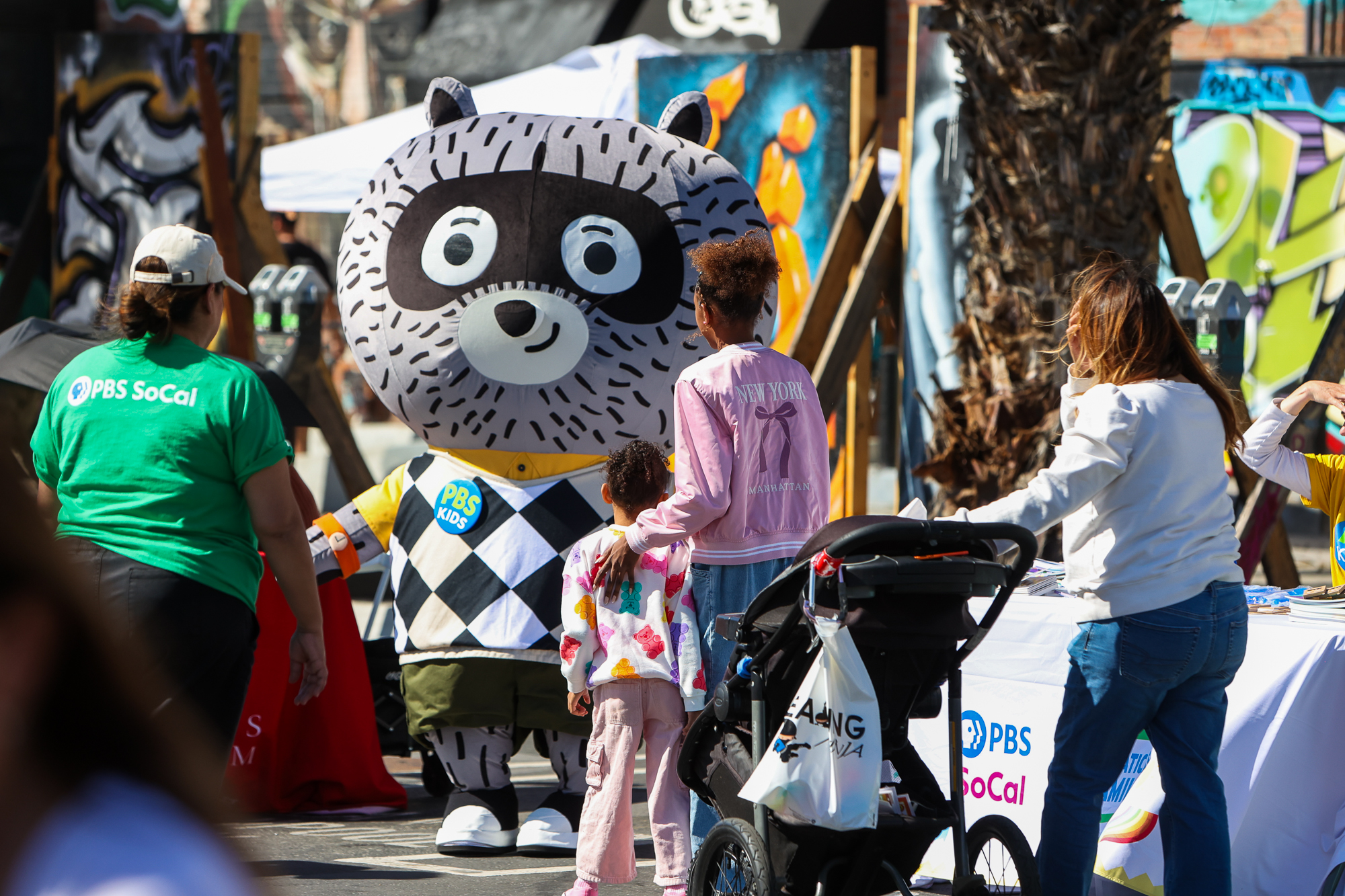 A costumed PBS character, Carl the Collector is seen engaging with children and adults. Carl is a raccoon with a large mascot style head. He is wearing a yellow short-sleeved button-up shirt under a black-and-white argyle sweater vest, green cargo shorts and a PBS Kids button. One adult with a child and another adult with a stroller are approaching Carl as he holds his arms wide open.