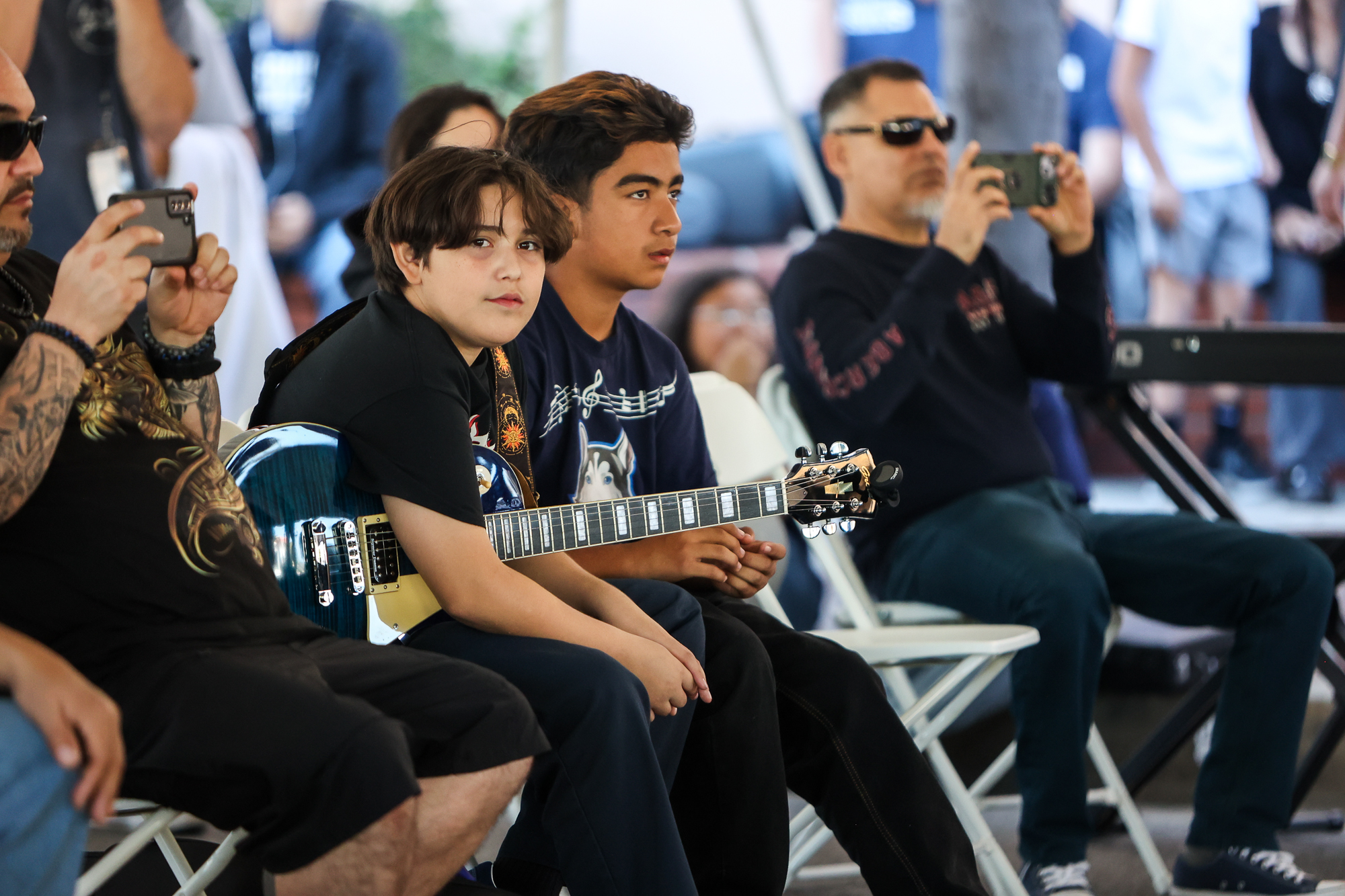Two elementary students sit in the audience, waiting to take the stage. One boy is holding his guitar and parents are scene recording the performances.