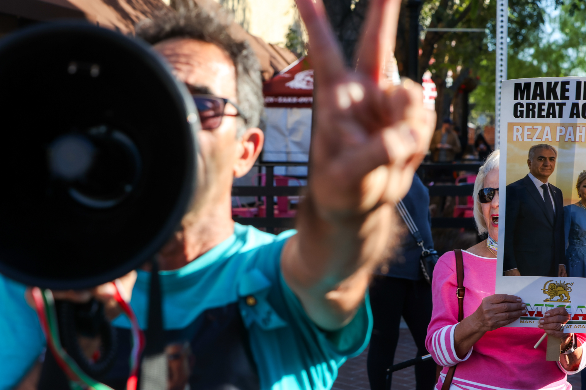 A blurred image of a man speaking into a megaphone and making a peace sign and a woman holding a sign in support of Iran.
