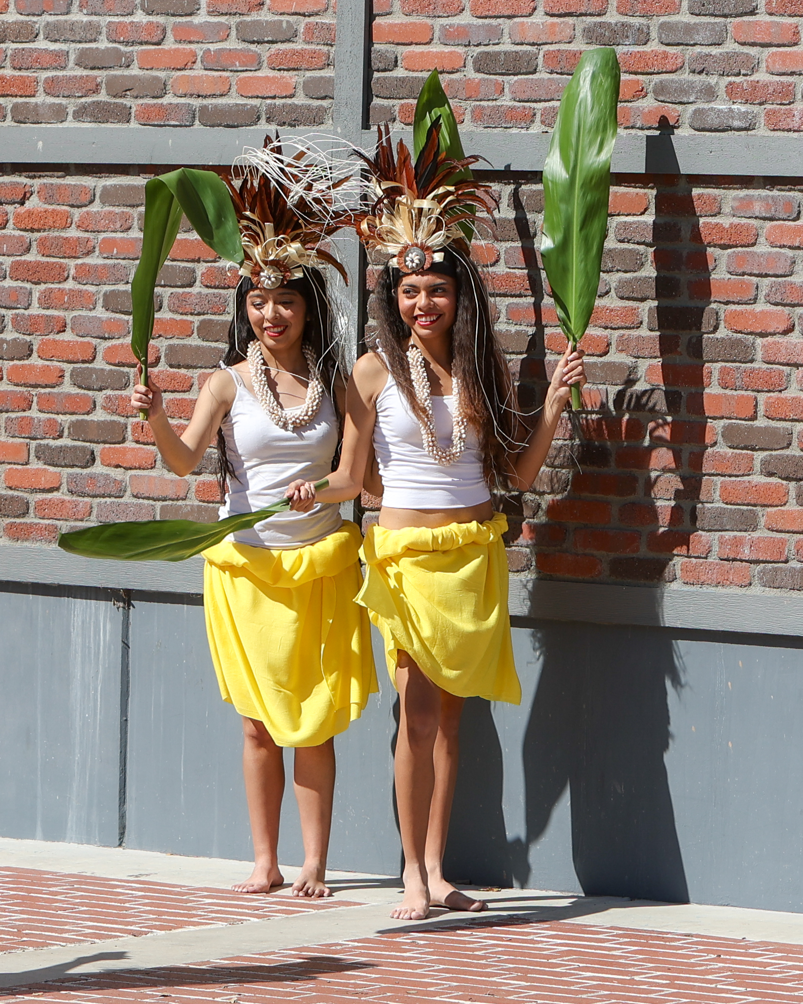 Two barefoot dancers in yellow wrap skirts and white tank tops hold banana leaves and wear elaborate headpieces.