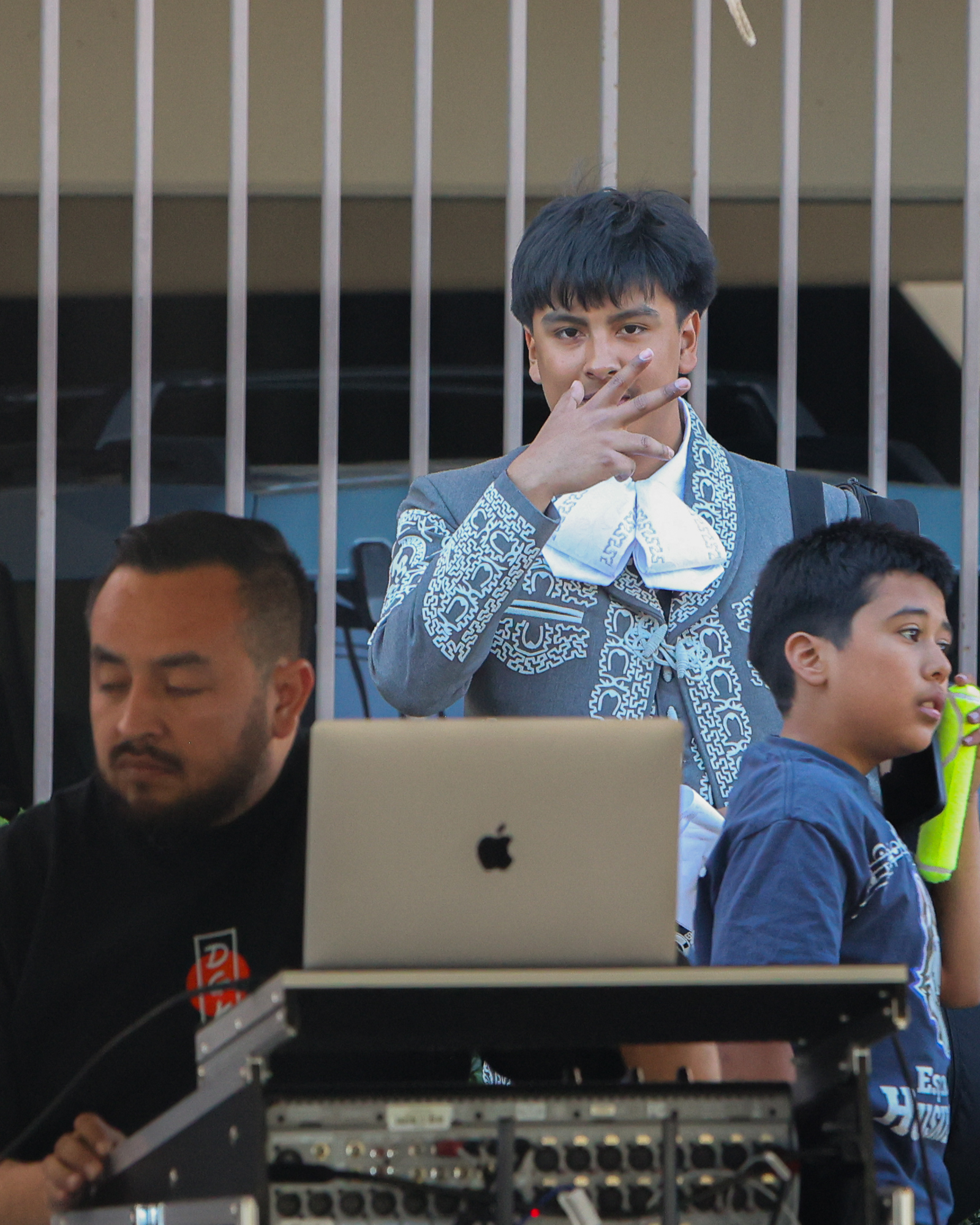A teenager dressed in a blue-grey mariachi suit stands behind a stage crew member using a Macbook for the production. A younger child passes by.