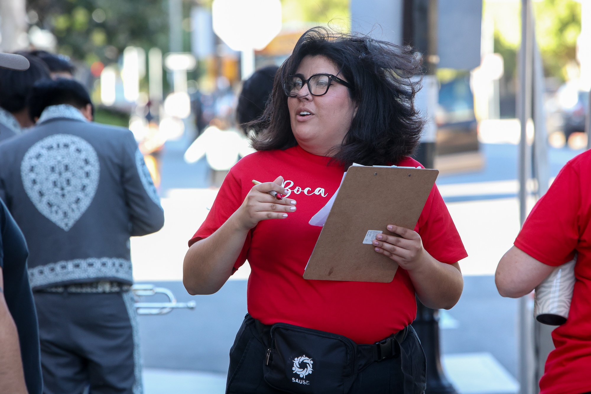 An event staffer has her clipboard and pen ready as her hair blows in the wind. She is wearing glasses, a red event shirt and a SAUSD fanny pack.