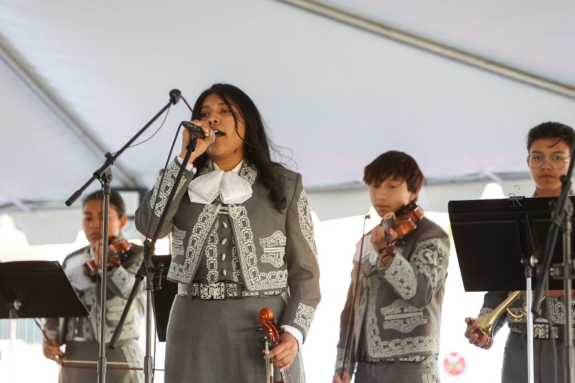 Four high school age mariachi band members wear traditional gray embroidered charro suits. The female lead singer is holding the mic as she performs.