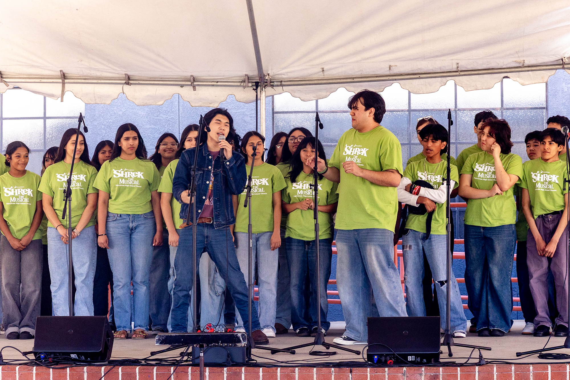 High school students, dressed in jeans and green "Shrek the Musical" shirts, perform on stage.