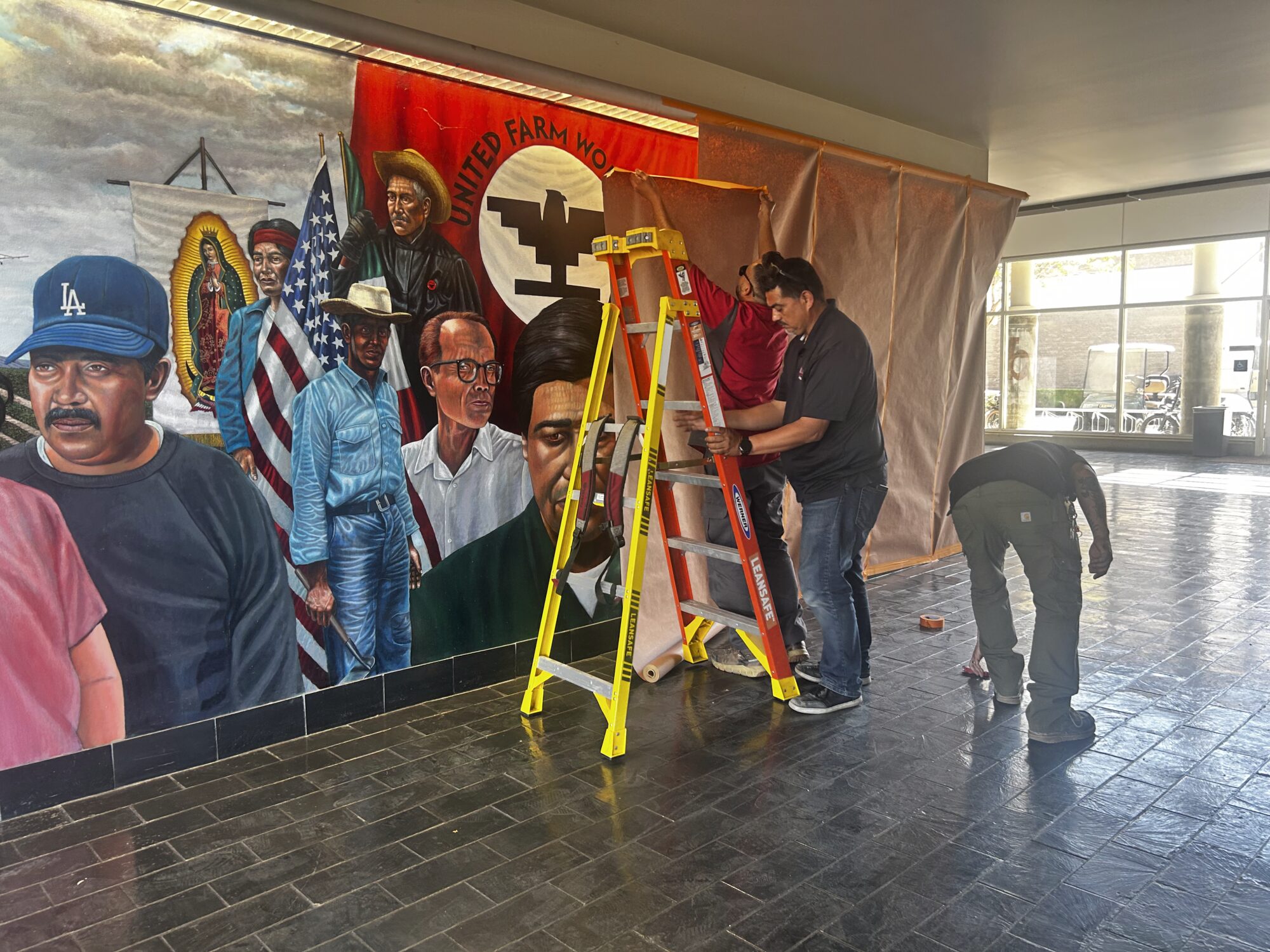 Three maintenece workers cover a colorful mural of Cesar Chavez and farmworkers on display in Santa Ana College.