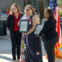 Two women holding plaques along with the presenter at an outdoor ceremony.