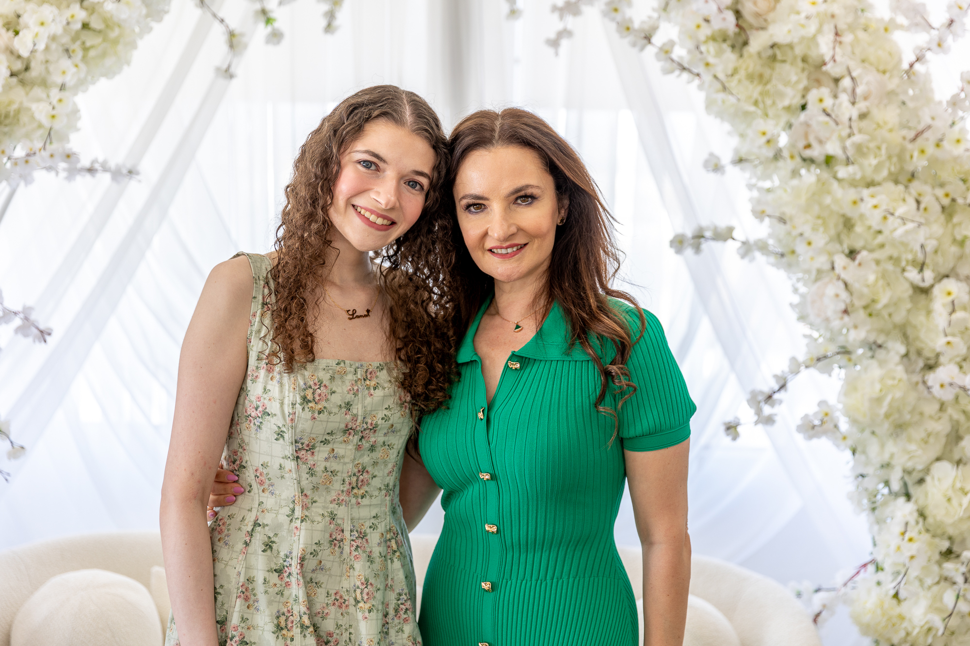 A young woman in a floral dress poses with her mother wearing a green dress. There is an arch of white flowers and sheer fabric in the background.