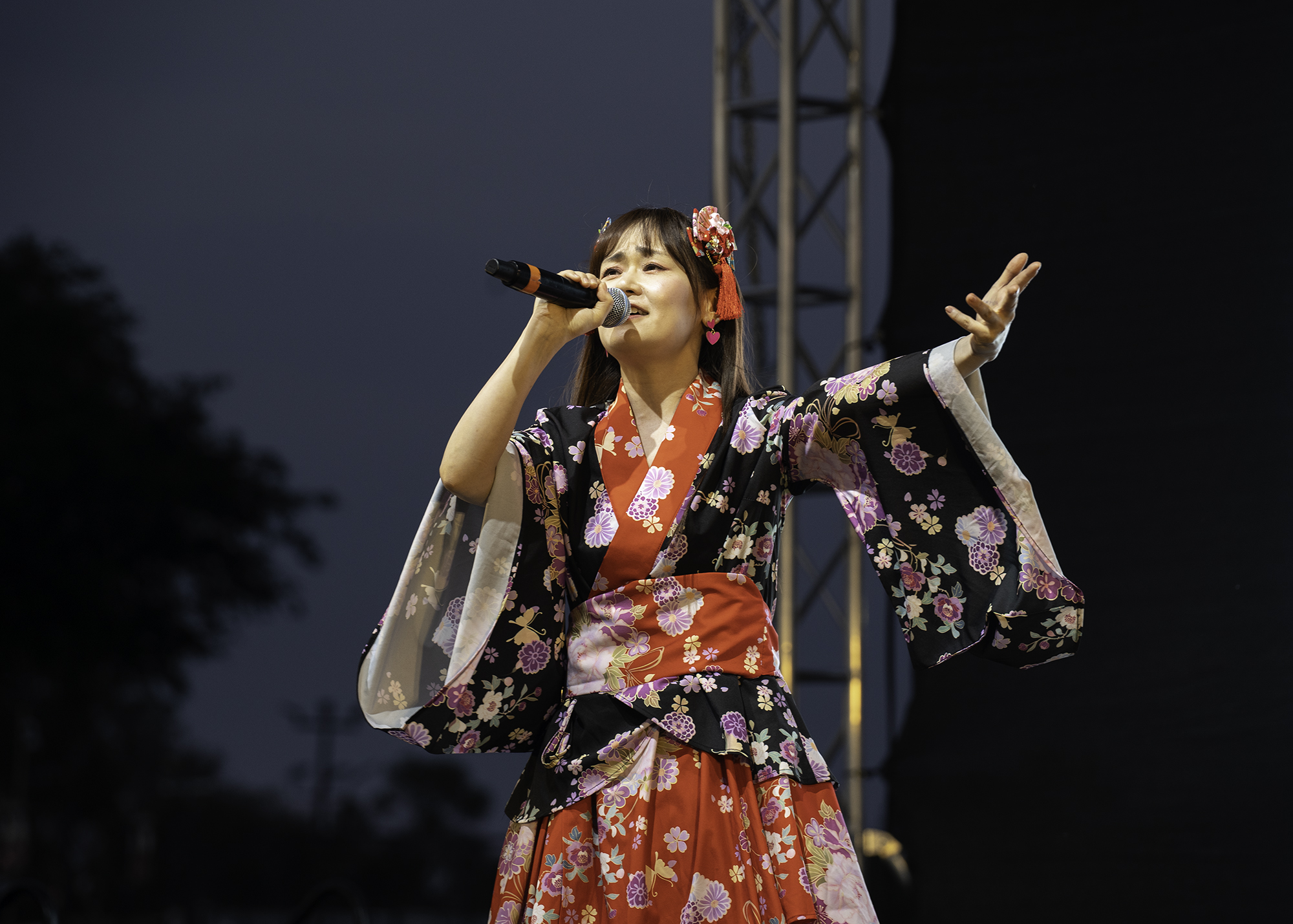 Female singer wears a floral Japanese kimono featuring reds and purples while holding a microphone.