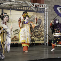 Three costumed individuals dance on stage holding umbrellas and wearing traditional Japanese kimonos.