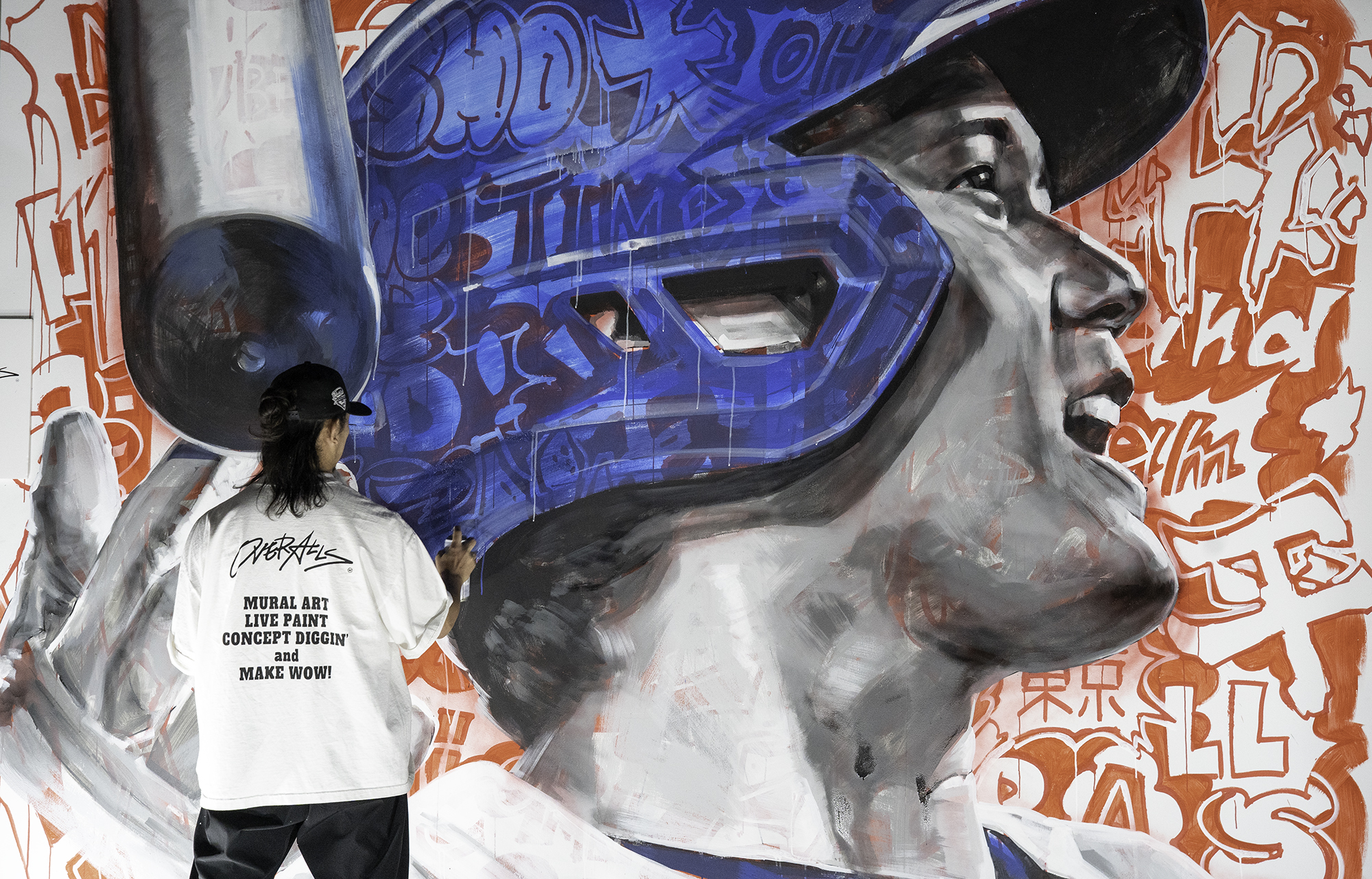 Male muralist with his back to the camera and holding a spray paint can works on a mural of baseball great Shohei Ohtani.