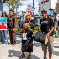Many protesters stand on a corner for the No Kings rally. A man and woman dressed in black use percussion instruments two protesters wearing masks stand behind them to the right holding signs.