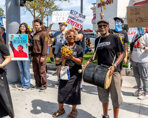 Many protesters stand on a corner for the No Kings rally. A man and woman dressed in black use percussion instruments two protesters wearing masks stand behind them to the right holding signs.
