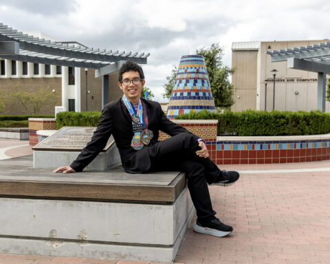 Male college student dressed in black suit and dress shirt wearing various colorful marathon medals. He sits on concrete platform in front of a colorful tile fountain.