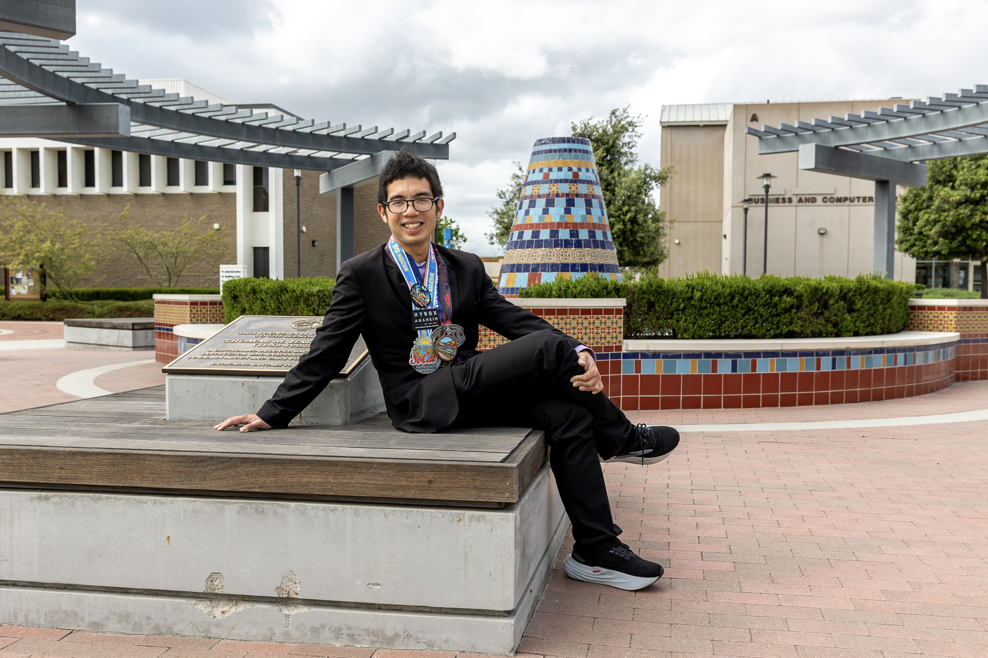 Male college student dressed in black suit and dress shirt wearing various colorful marathon medals. He sits on concrete platform in front of a colorful tile fountain.