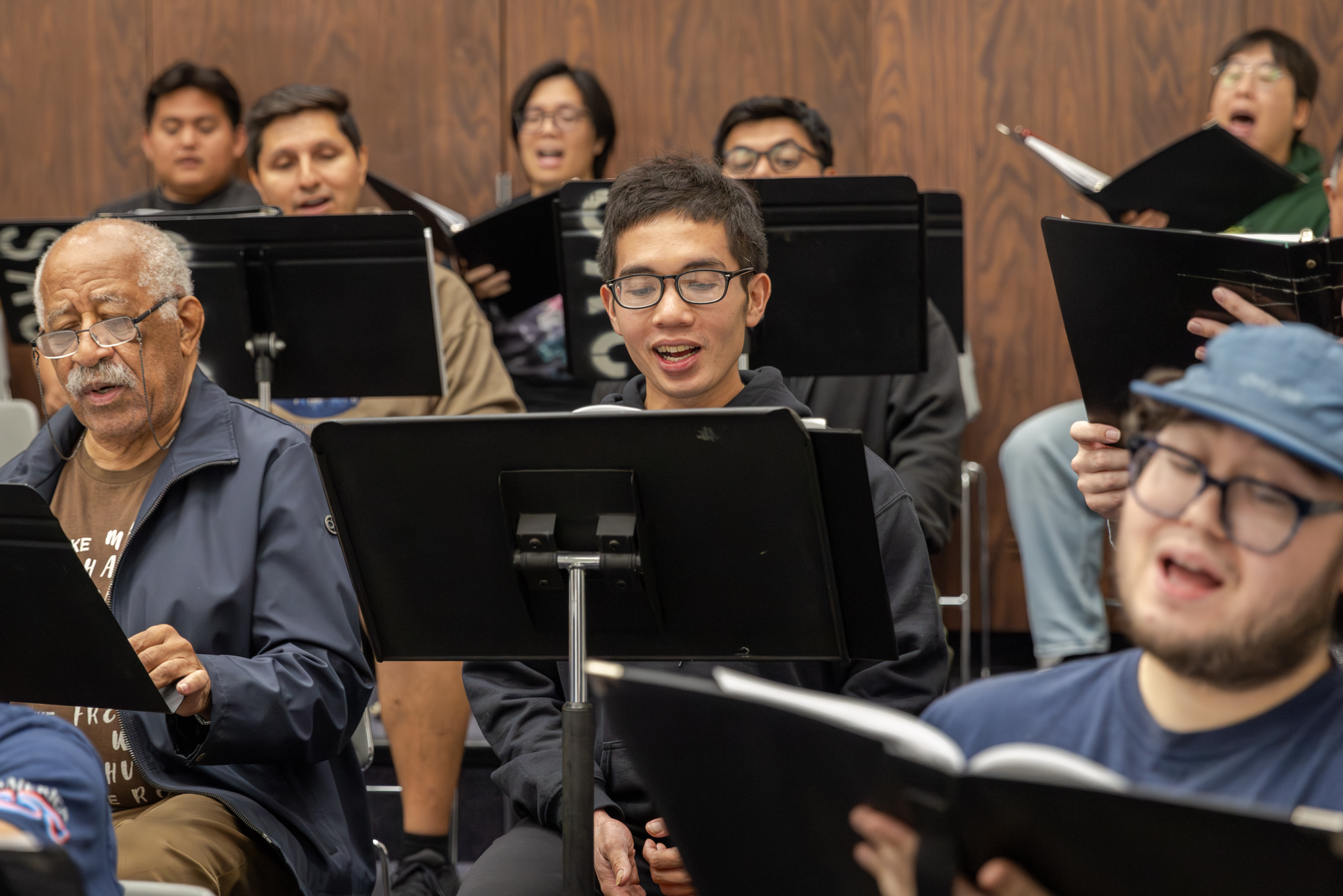 Men singing from sheet music placed on stands in a paneled rehearsal room.